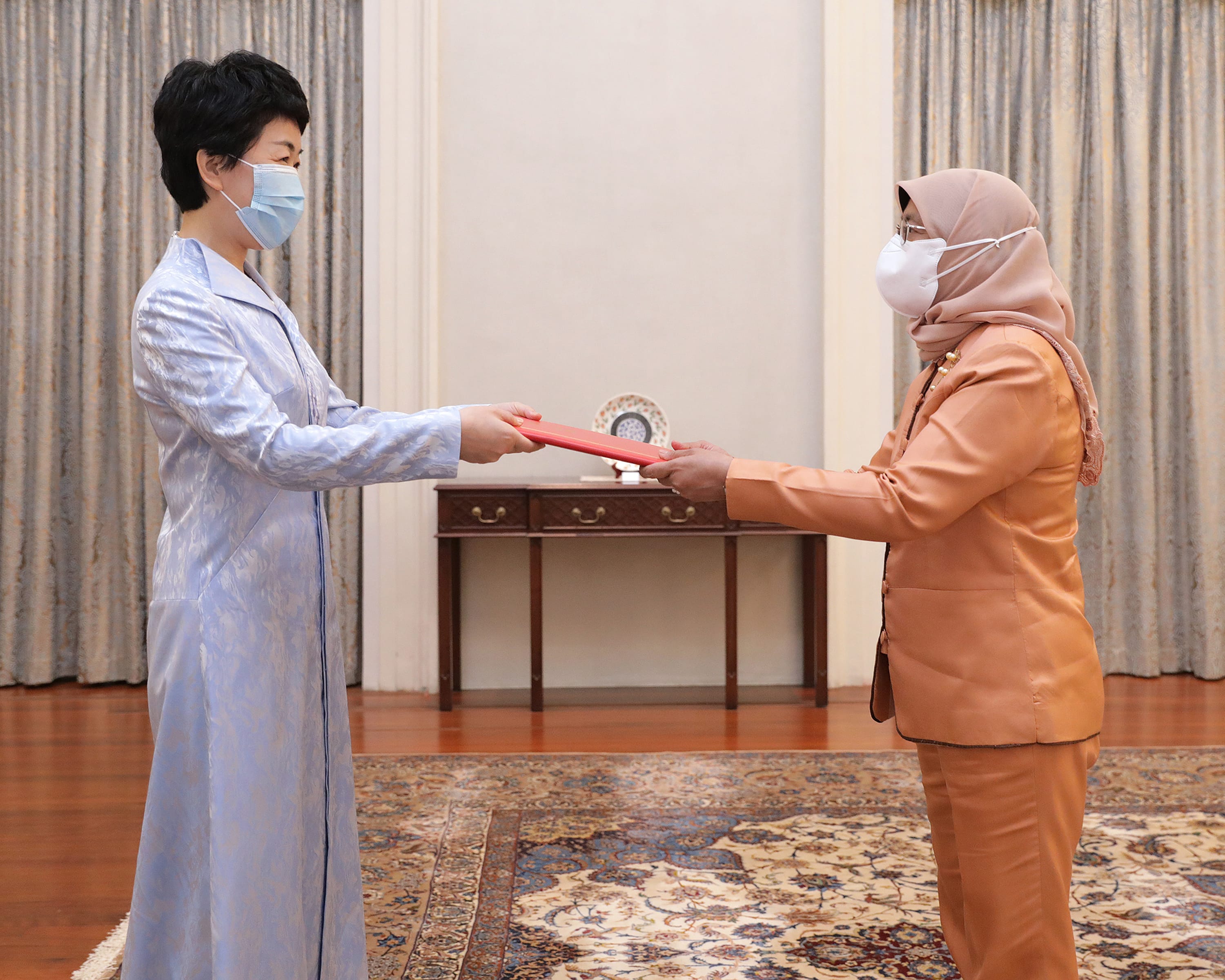 Two masked women exchanging a red document. One wears a blue dress, the other a brown suit and hijab.